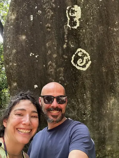 Two people smiling in front of a lichen-covered sandstone boulder at Savassona